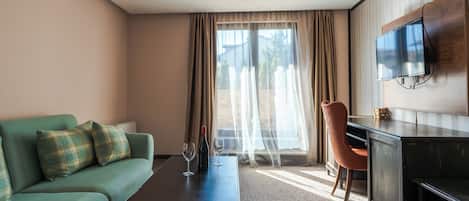 Une chambre d’hôtel avec un canapé vert, un bureau en bois, un téléviseur fixé au mur et une fenêtre ornée de rideaux légers.