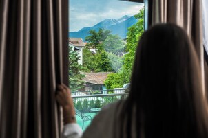 Una persona mirando por una ventana con cortinas, con vistas a un paisaje montañoso y un pueblo con edificios tradicionales.