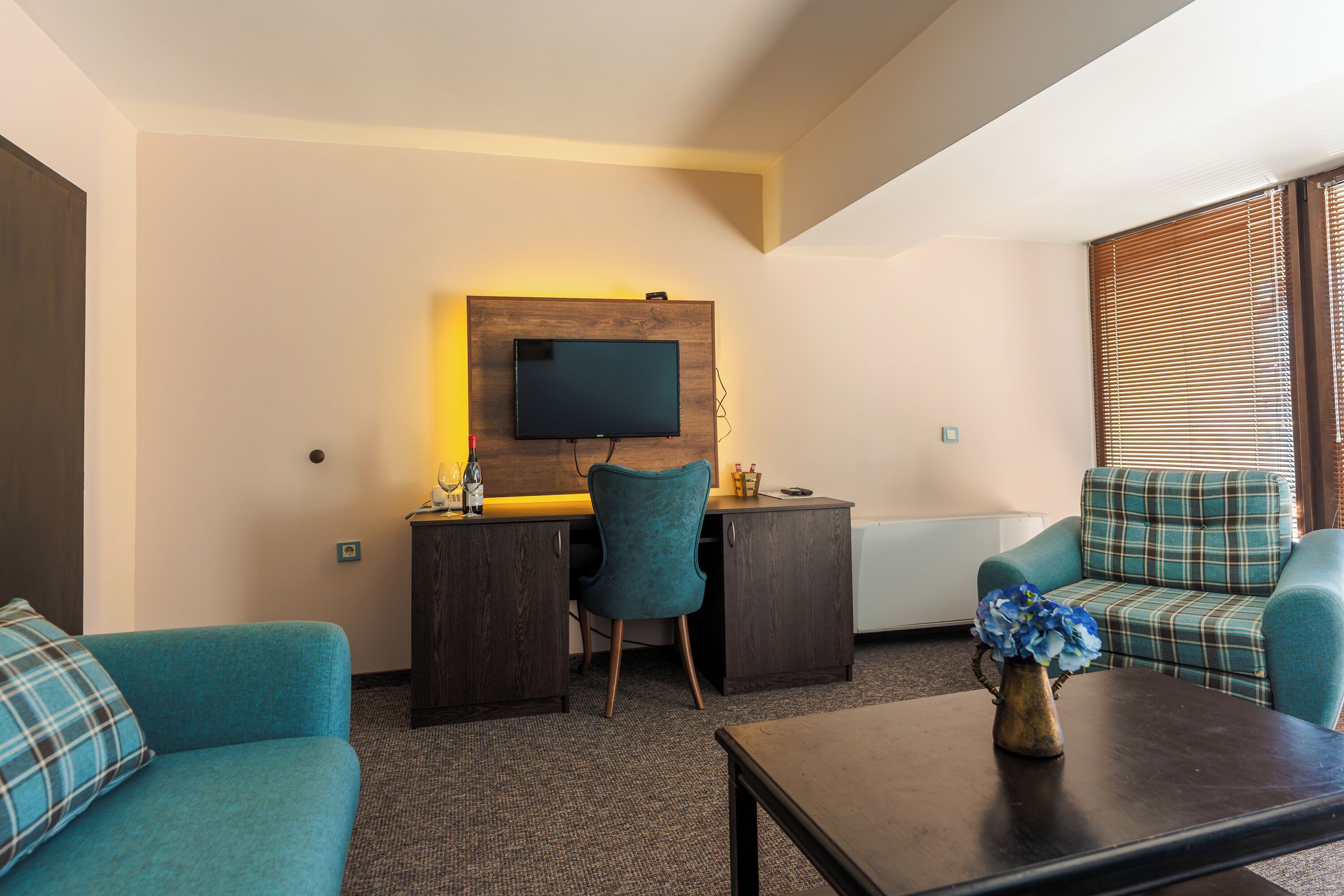 A modern hotel room with a flat-screen TV mounted on a wooden panel, a desk with a chair, a sofa, and a coffee table with a vase of flowers.