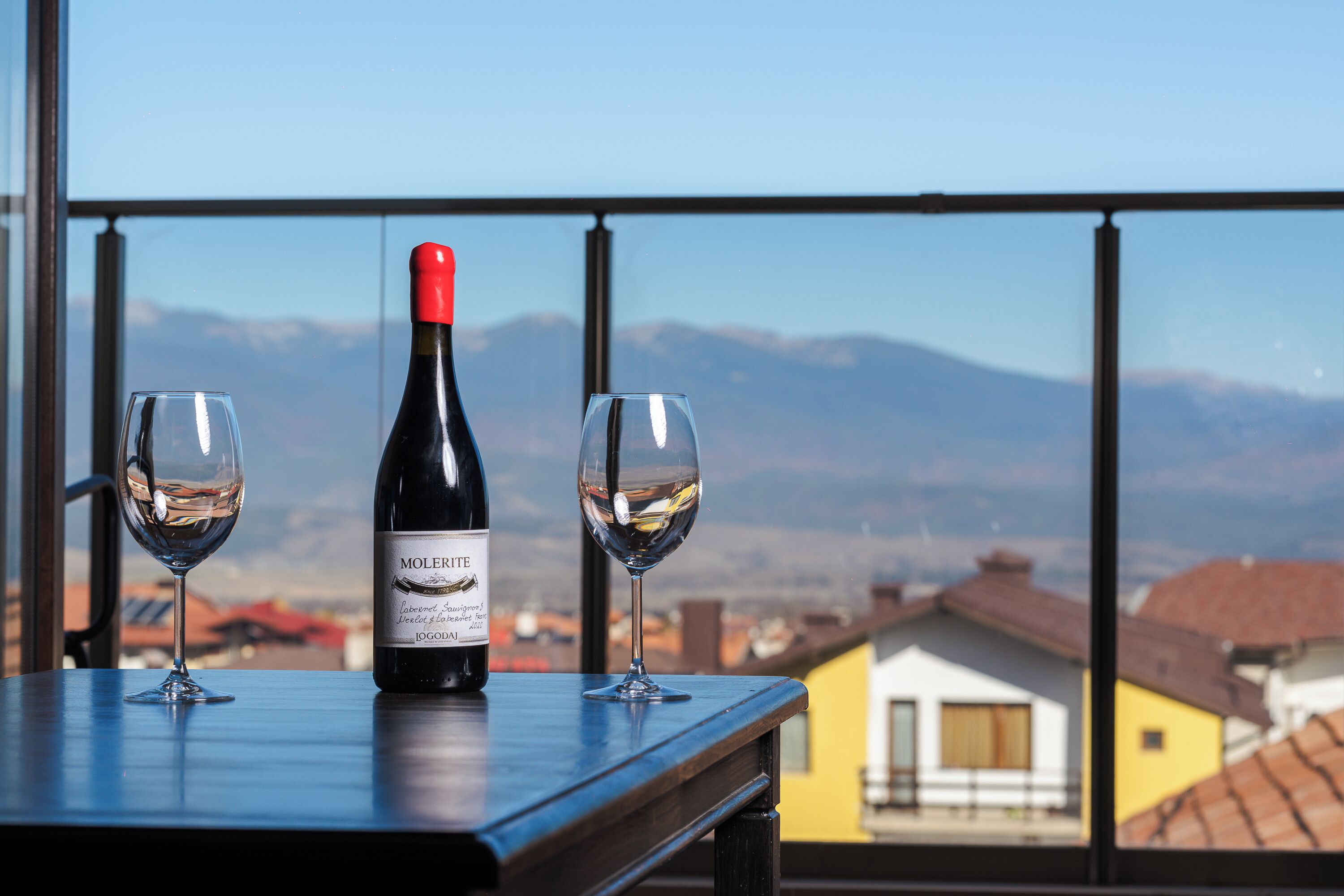 A bottle of wine and two glasses on a table with a view of mountains and buildings.