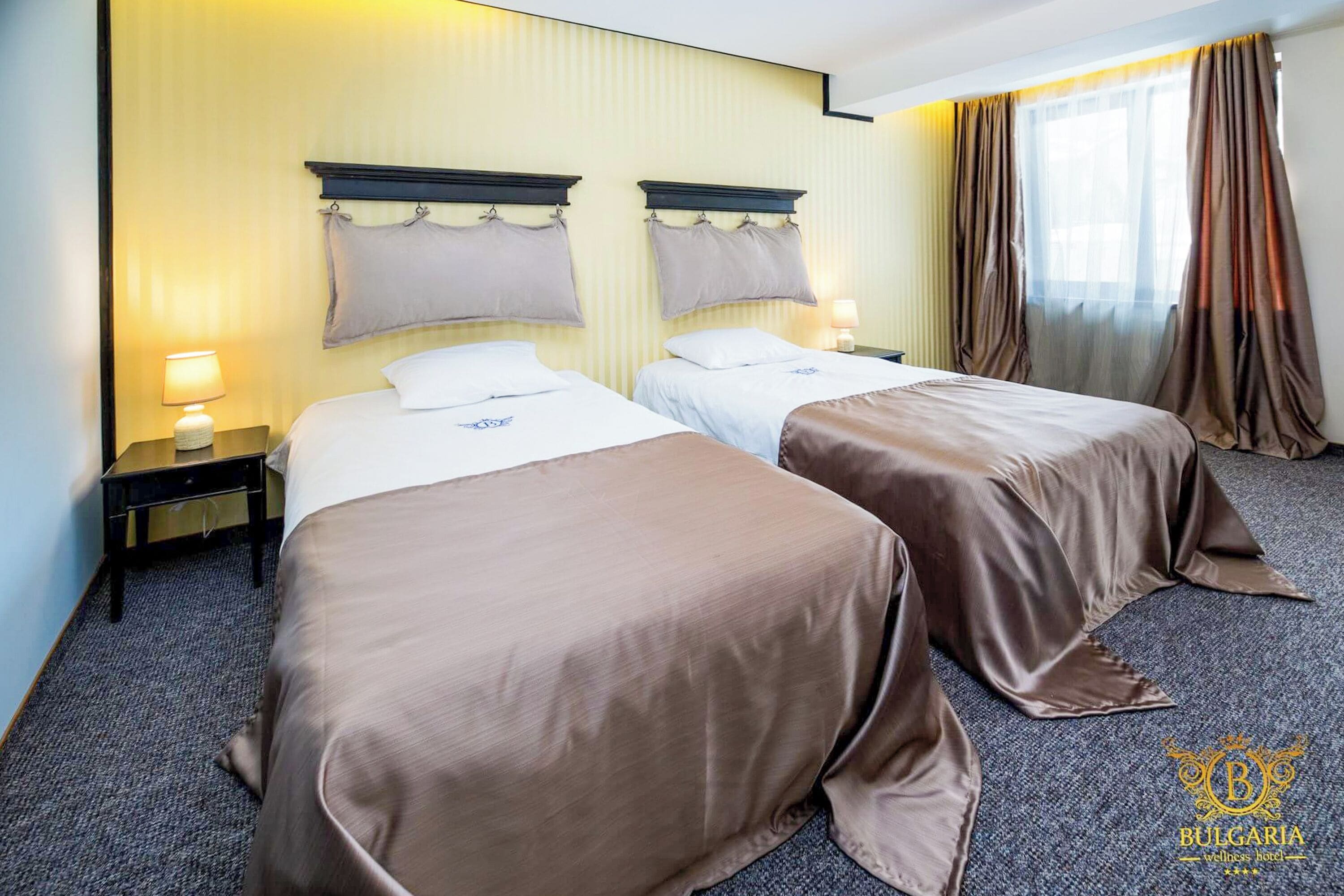A hotel room with two beds, a nightstand with a lamp, and a window with curtains.