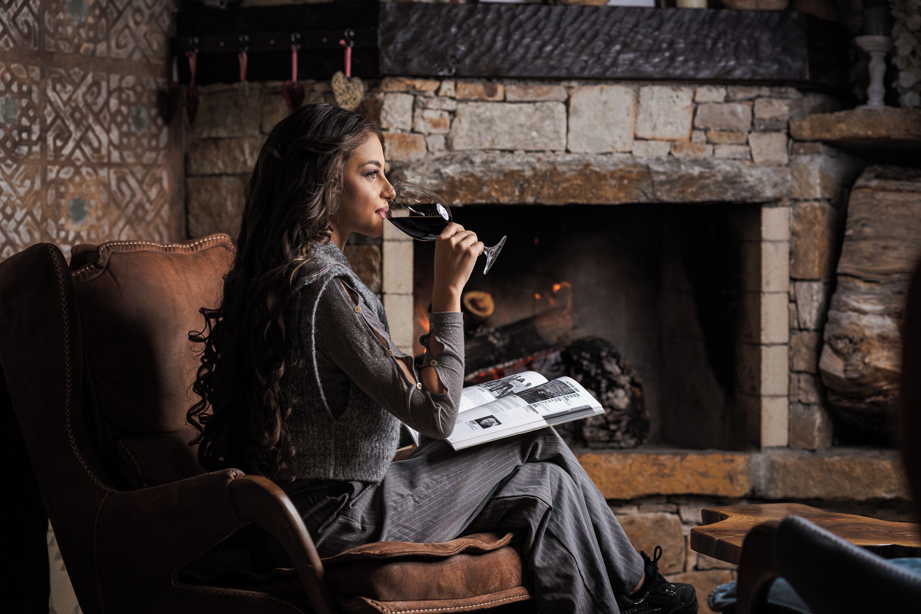 Una persona sentada junto a la chimenea, leyendo un libro y sosteniendo una copa de vino.