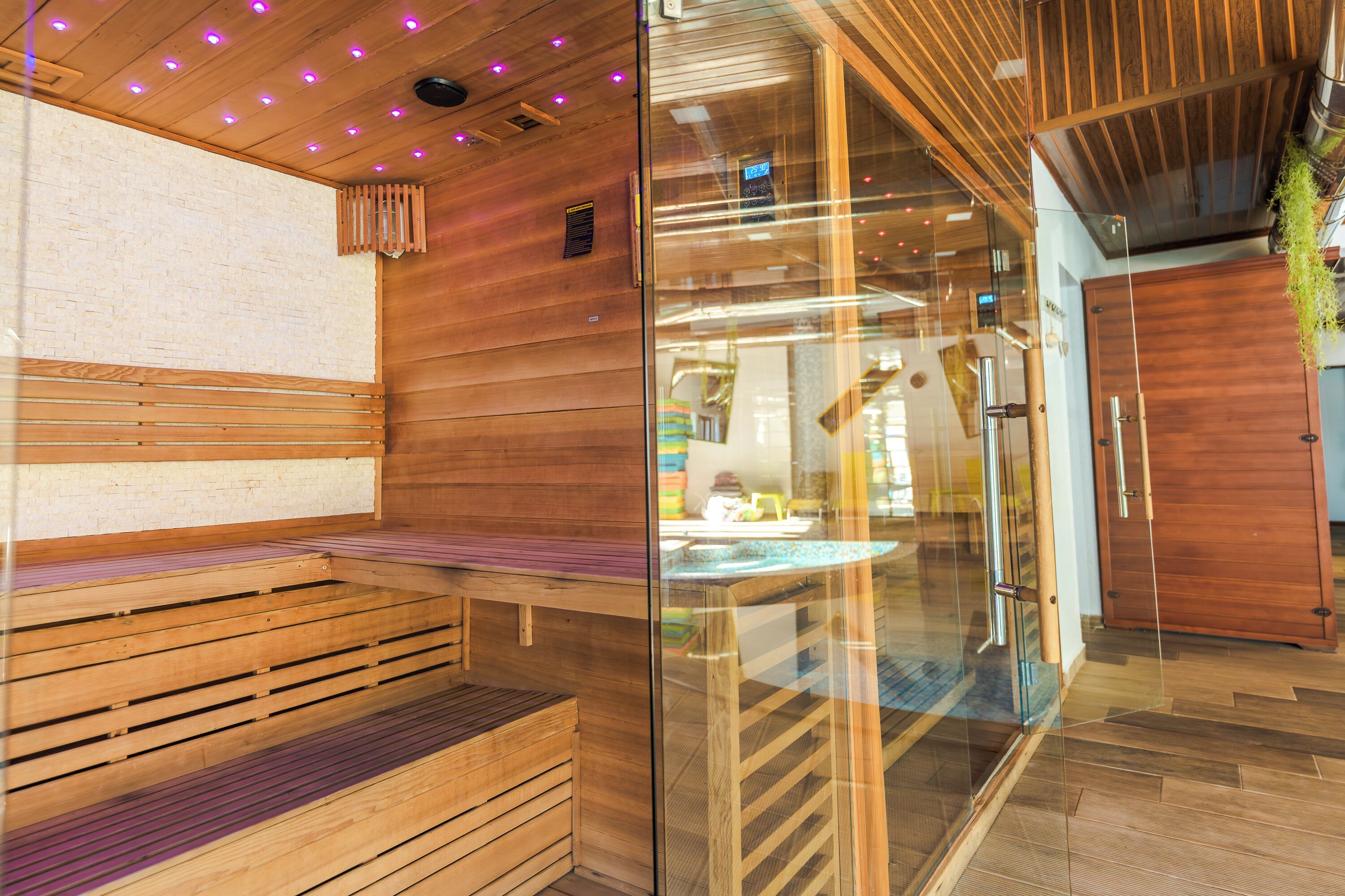 Een houten sauna met banken en een glazen deur, houten wanden en een plafond met paarse verlichting.