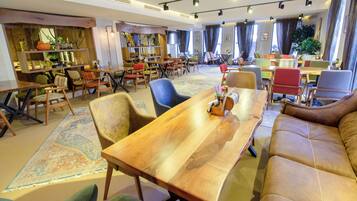 Un amplio vestíbulo de hotel con una mesa de madera en el centro, varias zonas de estar y un ventanal con cortinas.