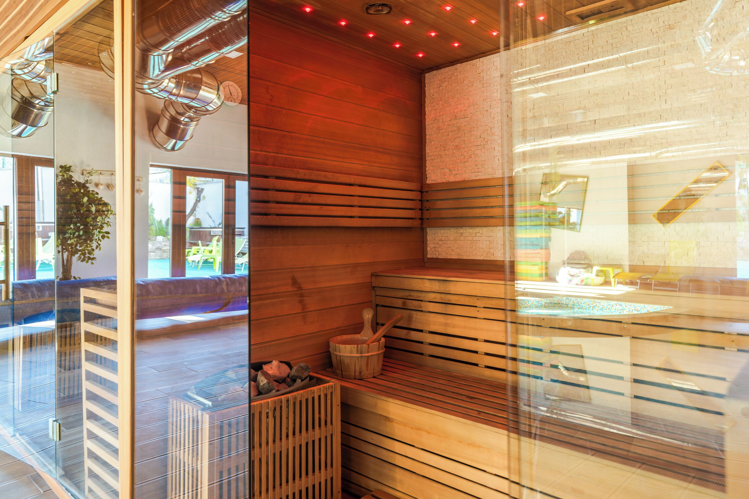 A sauna with wooden benches, a glass partition, and a visible outdoor area with plants and a ladder.