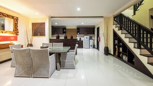 A spacious living area with a dining table and chairs, a kitchen in the background, and a staircase with a dark railing. - 2BR Private Pool Tiga Samudra Villa (badung)