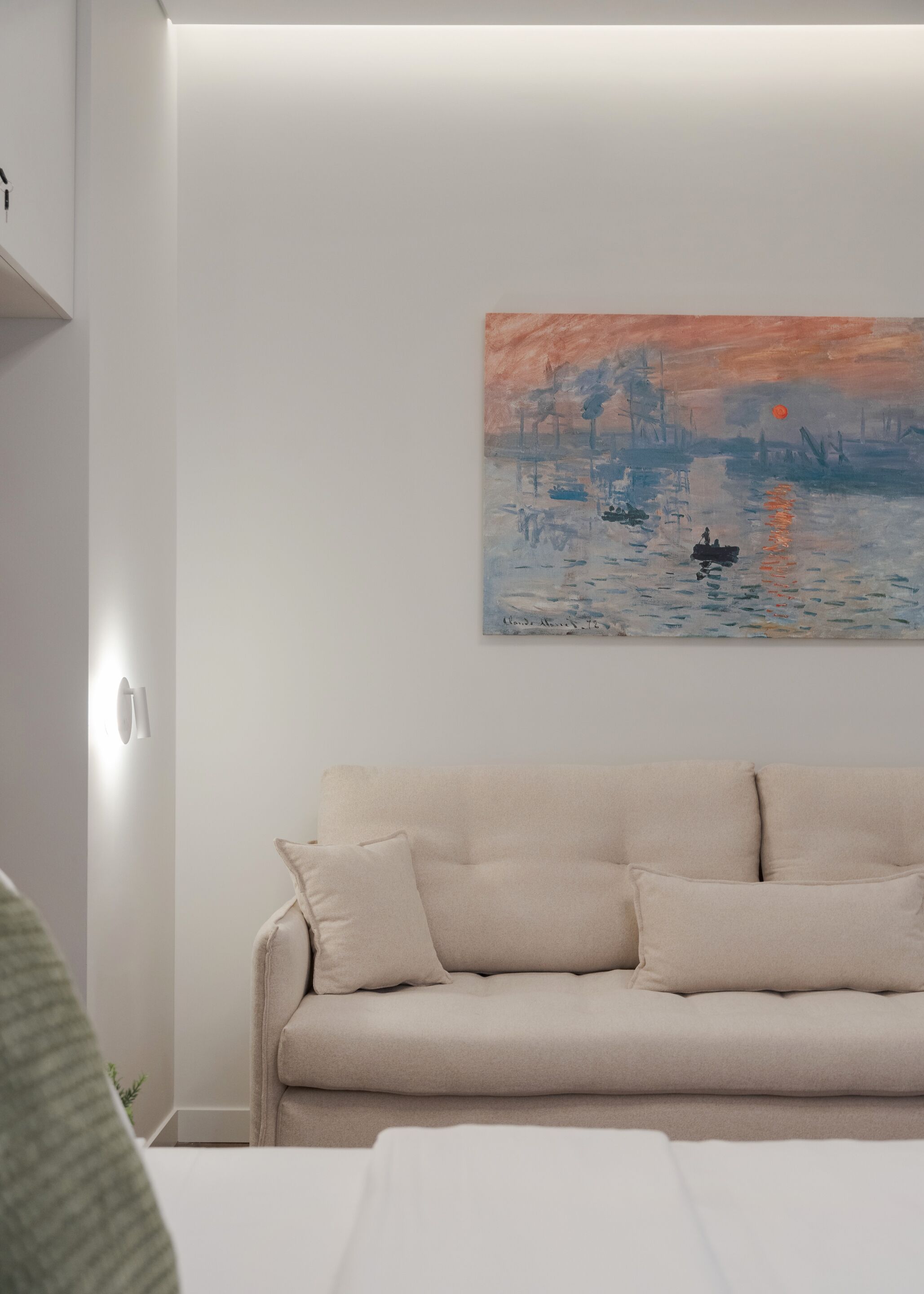 A room with a beige sofa, a painting of a sunset over water, and a white wall.