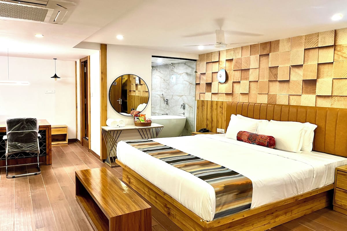 A modern hotel room with a wooden headboard, a large bed with striped bedding, a bedside table, a round mirror, a small desk with a chair, and a bench.