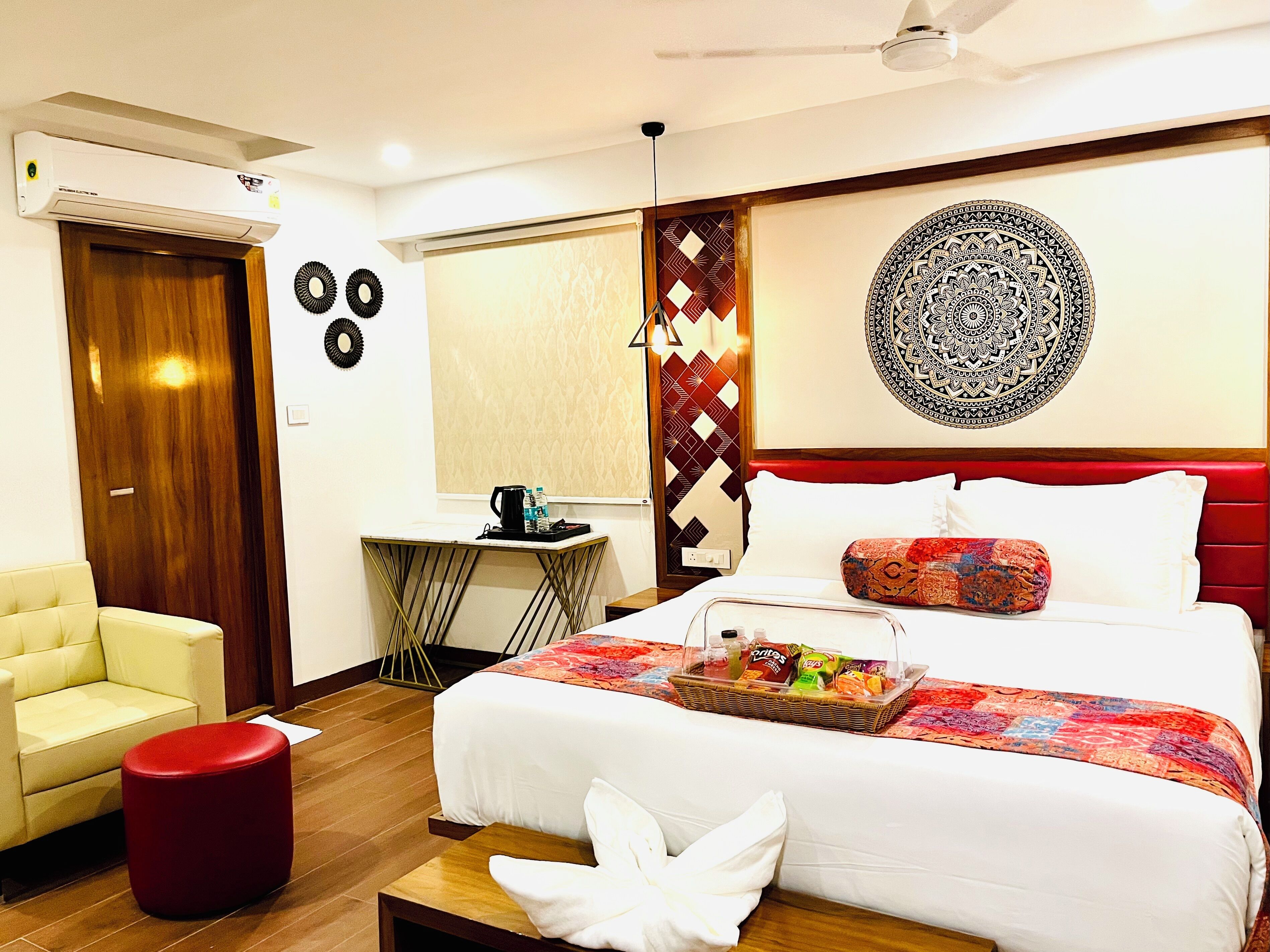 A hotel room with a bed, a desk, a chair, a red ottoman, and a decorative wall piece.