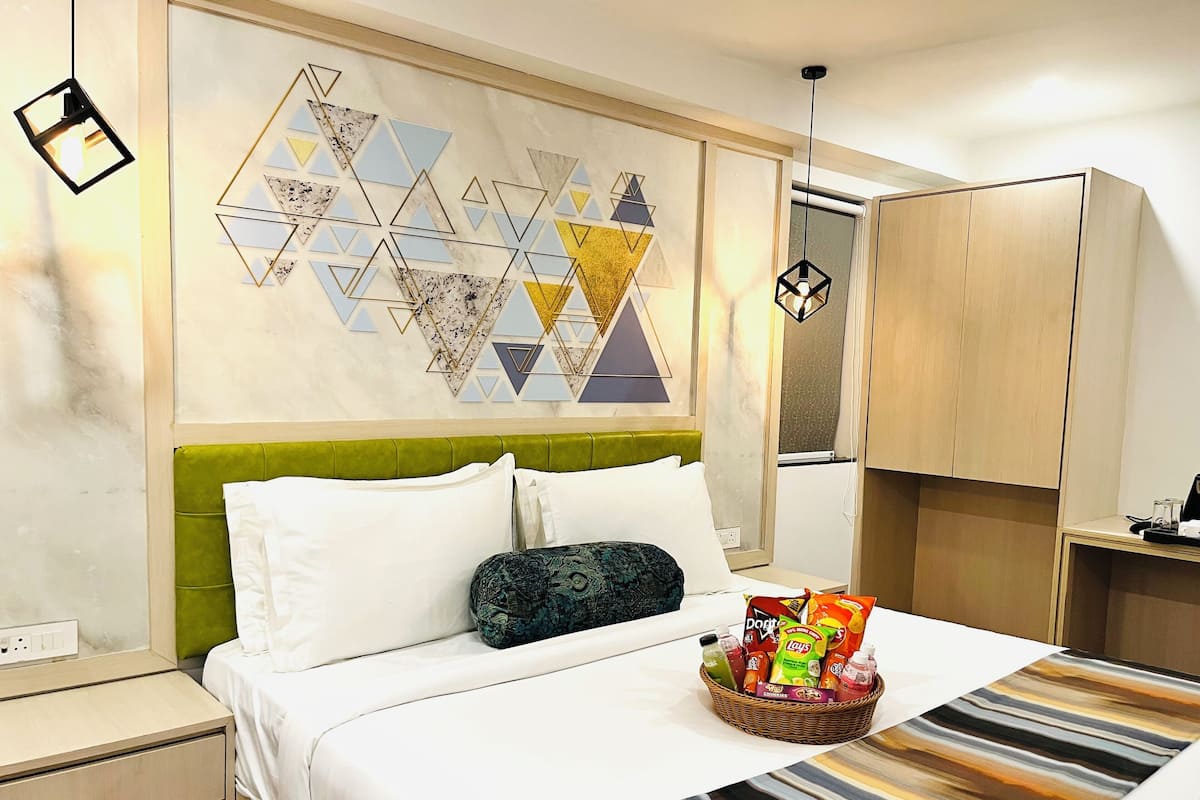 A hotel room with a bed, green headboard, white bedding, a wall art piece, a small table with a tray of snacks, and a wardrobe.