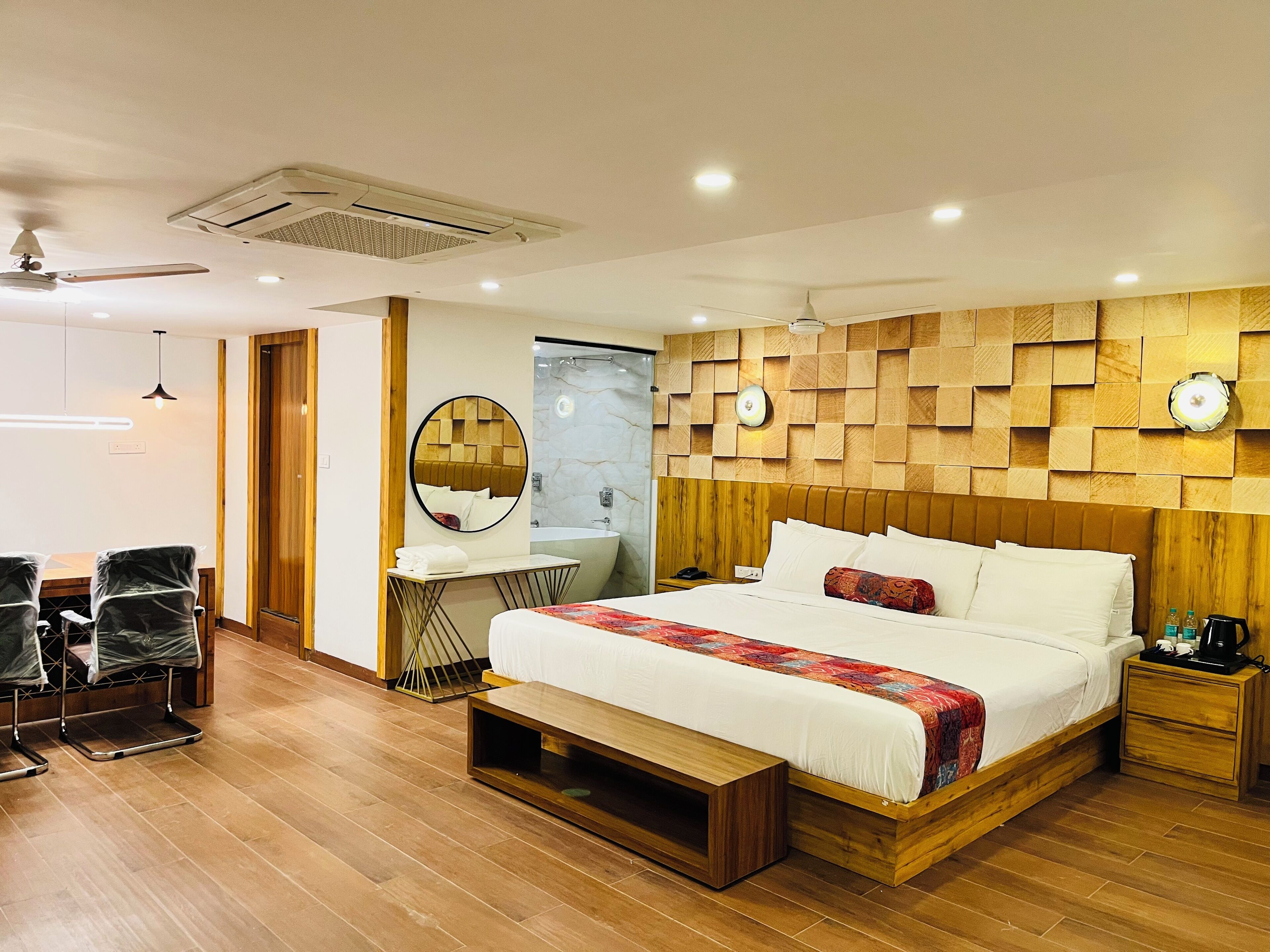 A modern bedroom with a large bed, wooden headboard, a round mirror, a vanity with a sink, and a desk with chairs.