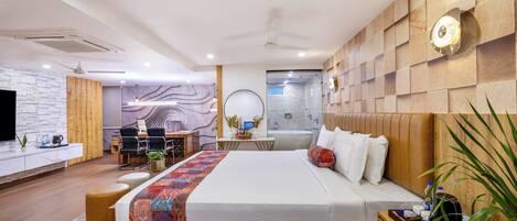 Una habitación de hotel moderna con cabecera de madera, una cama grande, un escritorio con silla, un baño pequeño y una zona de estar con sofá y mesa de centro.