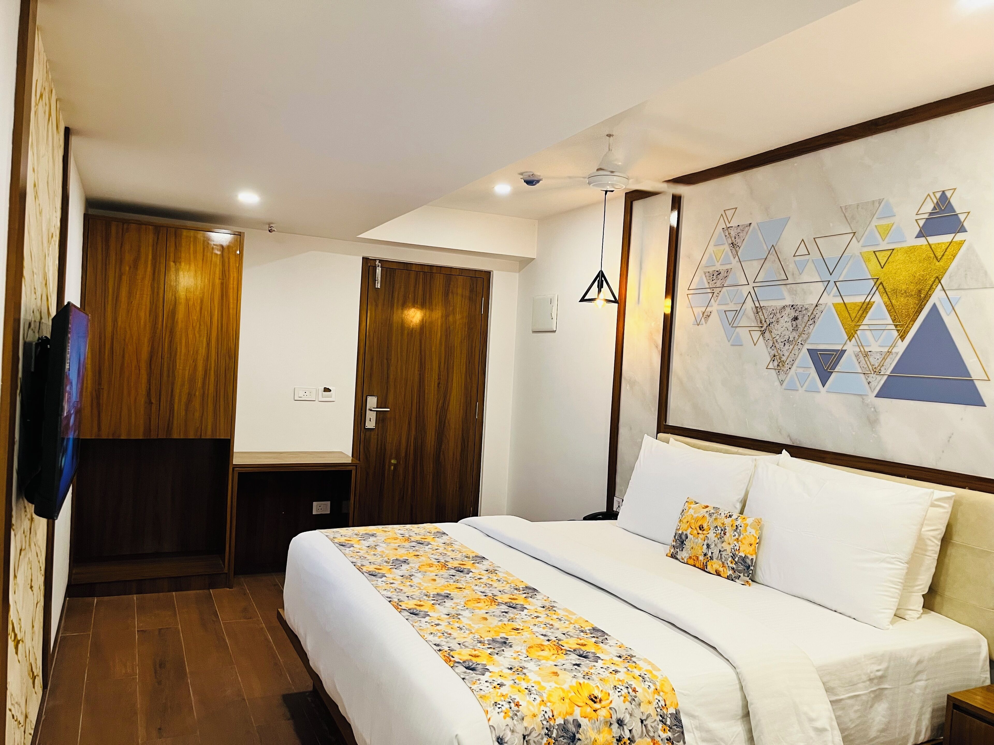 Une chambre d’hôtel équipée d’un lit, d’une armoire, d’un ventilateur de plafond et d’un mur orné d’œuvres d’art géométriques.
