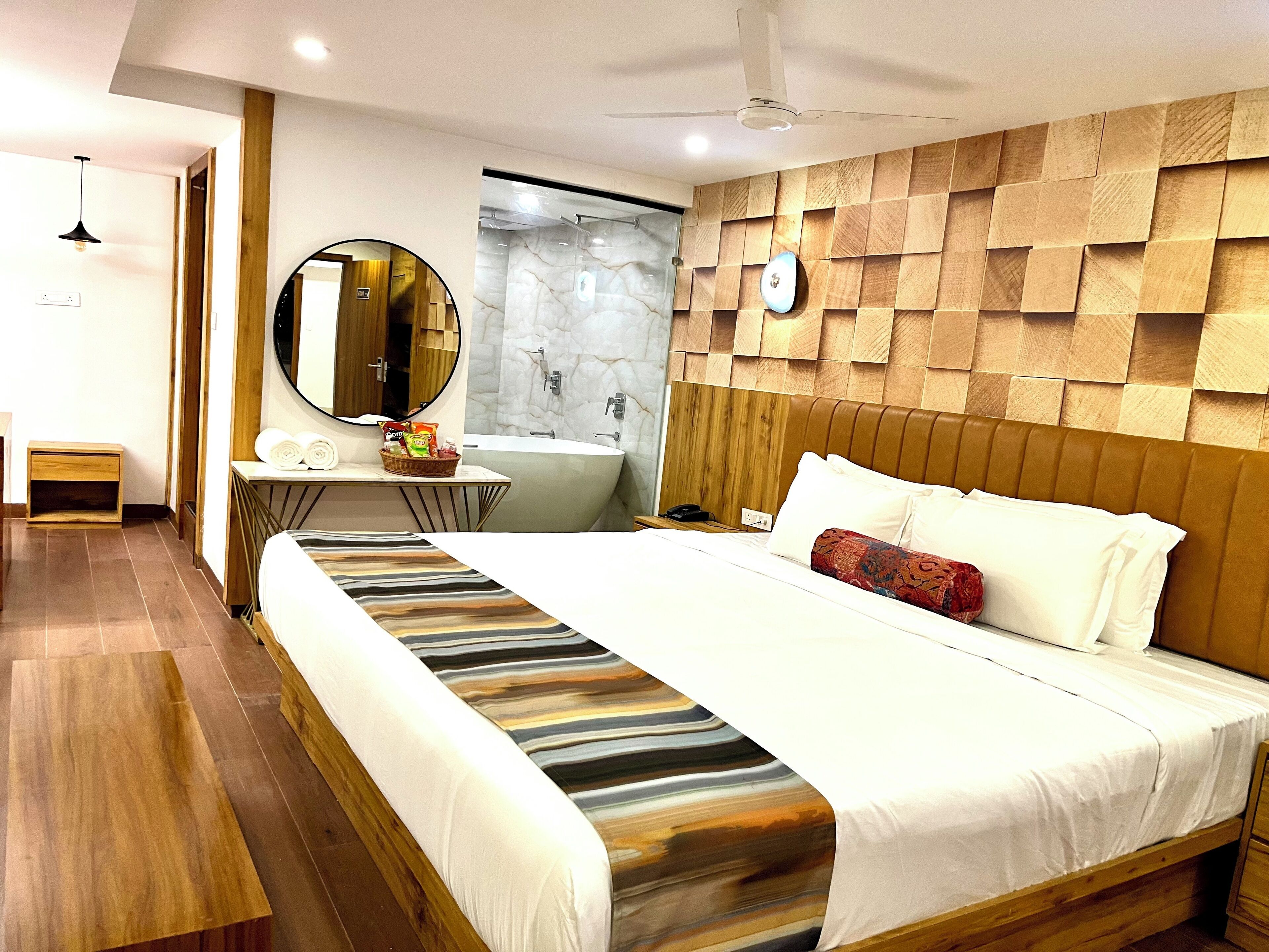 Una habitación de hotel moderna con una cama grande, cabecera de madera, un espejo redondo, una tocador con salpicadero de mármol y suelo de madera.