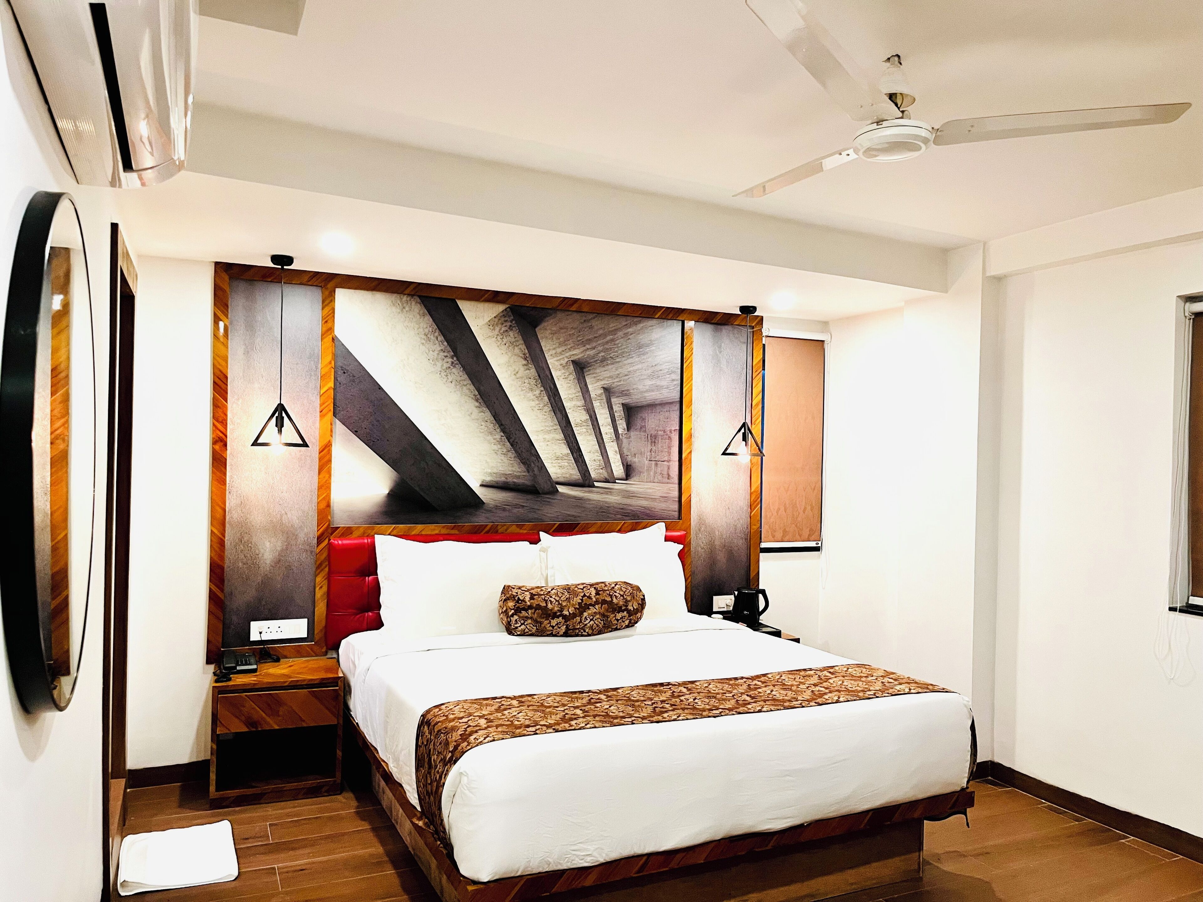 Habitación de hotel con una cama grande, cabecera de madera, cojín decorativo rojo, manta estampada, mesita de noche con lámpara y ventilador de techo.