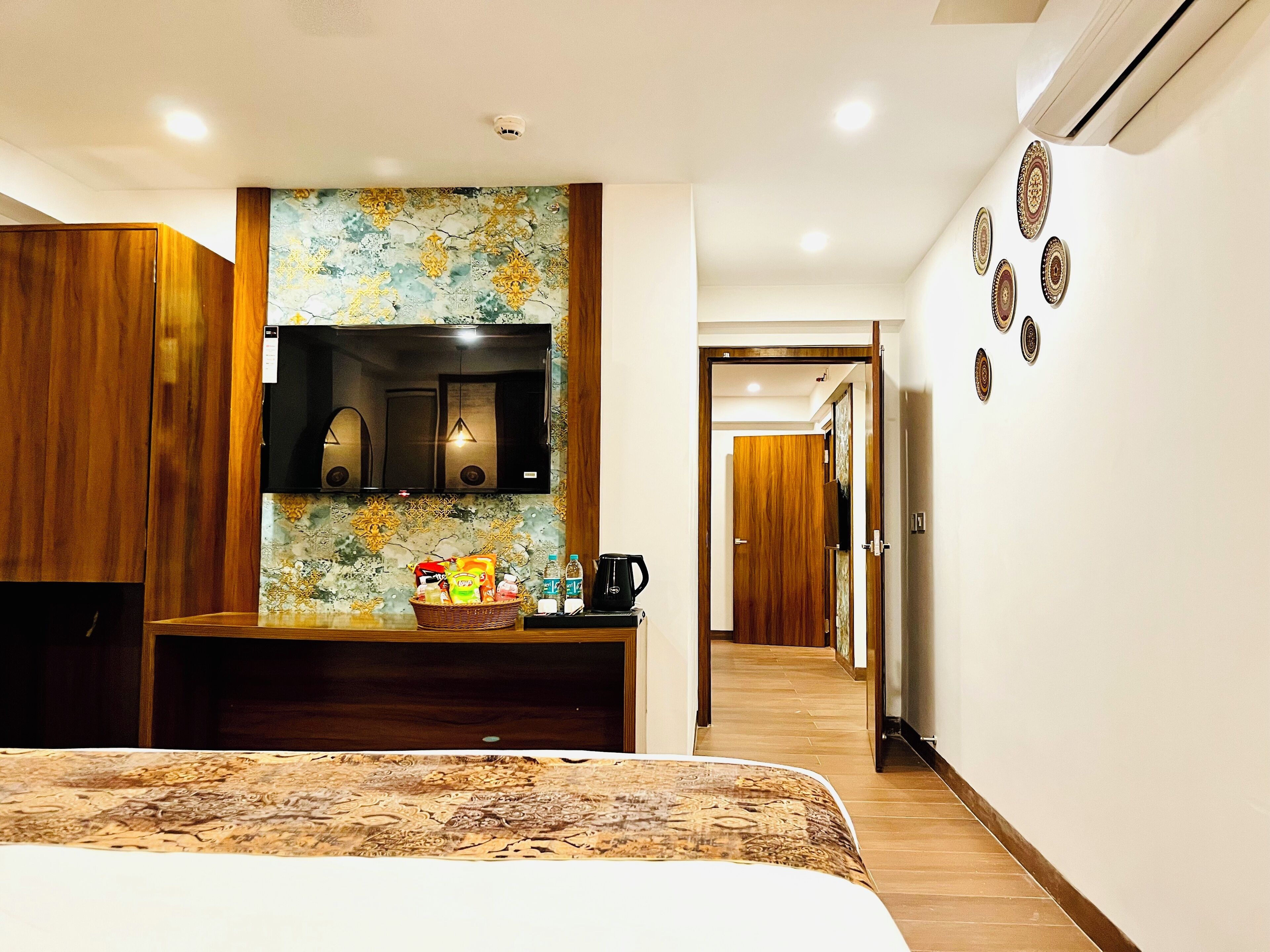 Habitación de hotel con cama, armario de madera, televisor, estante en la pared con aperitivos, cafetera y un pasillo que conduce a otra habitación.