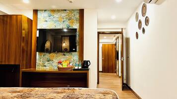 Habitación de hotel con cama, armario de madera, televisor, estante en la pared con aperitivos, cafetera y un pasillo que conduce a otra habitación.