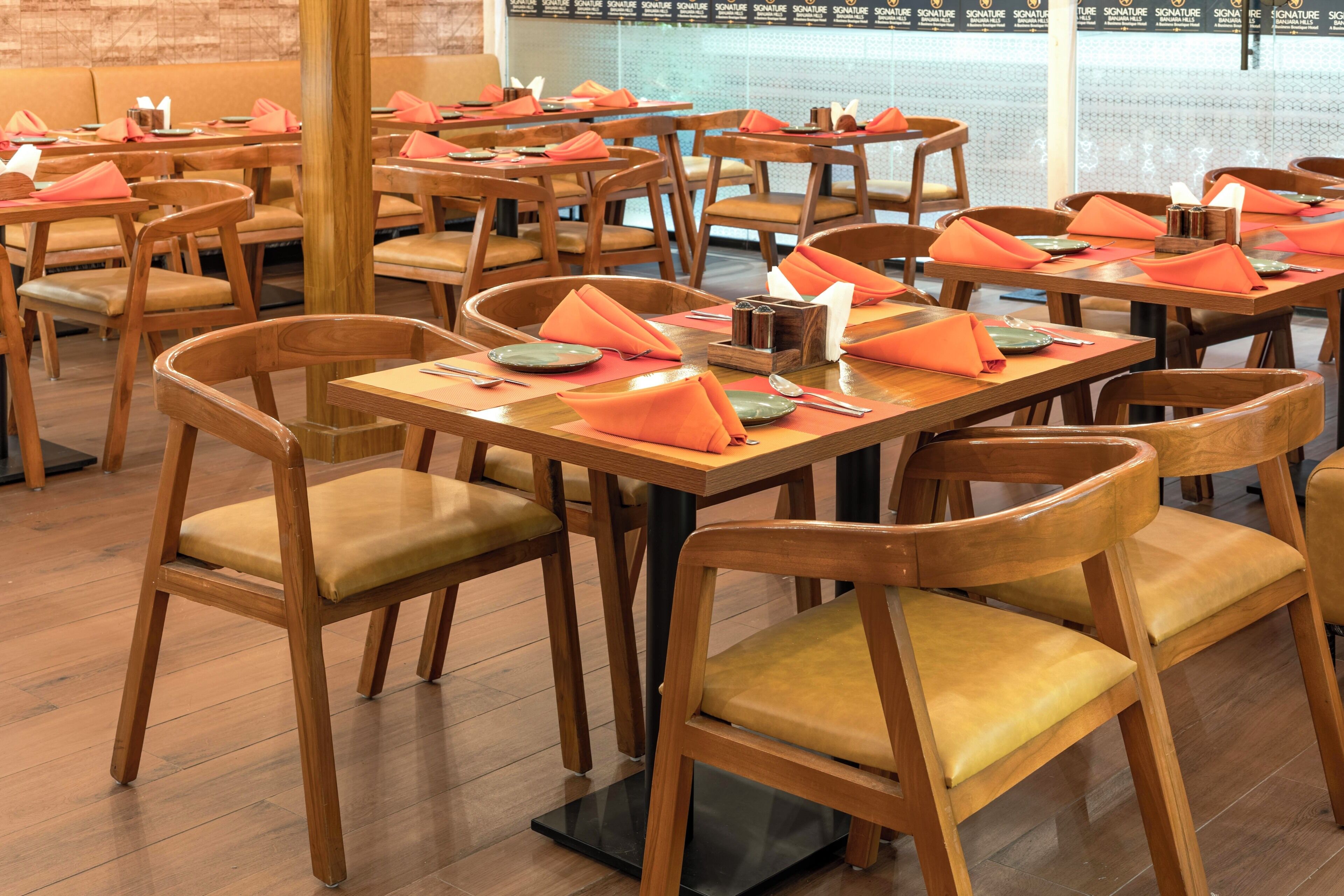 Interior de restaurante con mesas y sillas de madera, ordenado con servilletas anaranjadas y cubiertos.