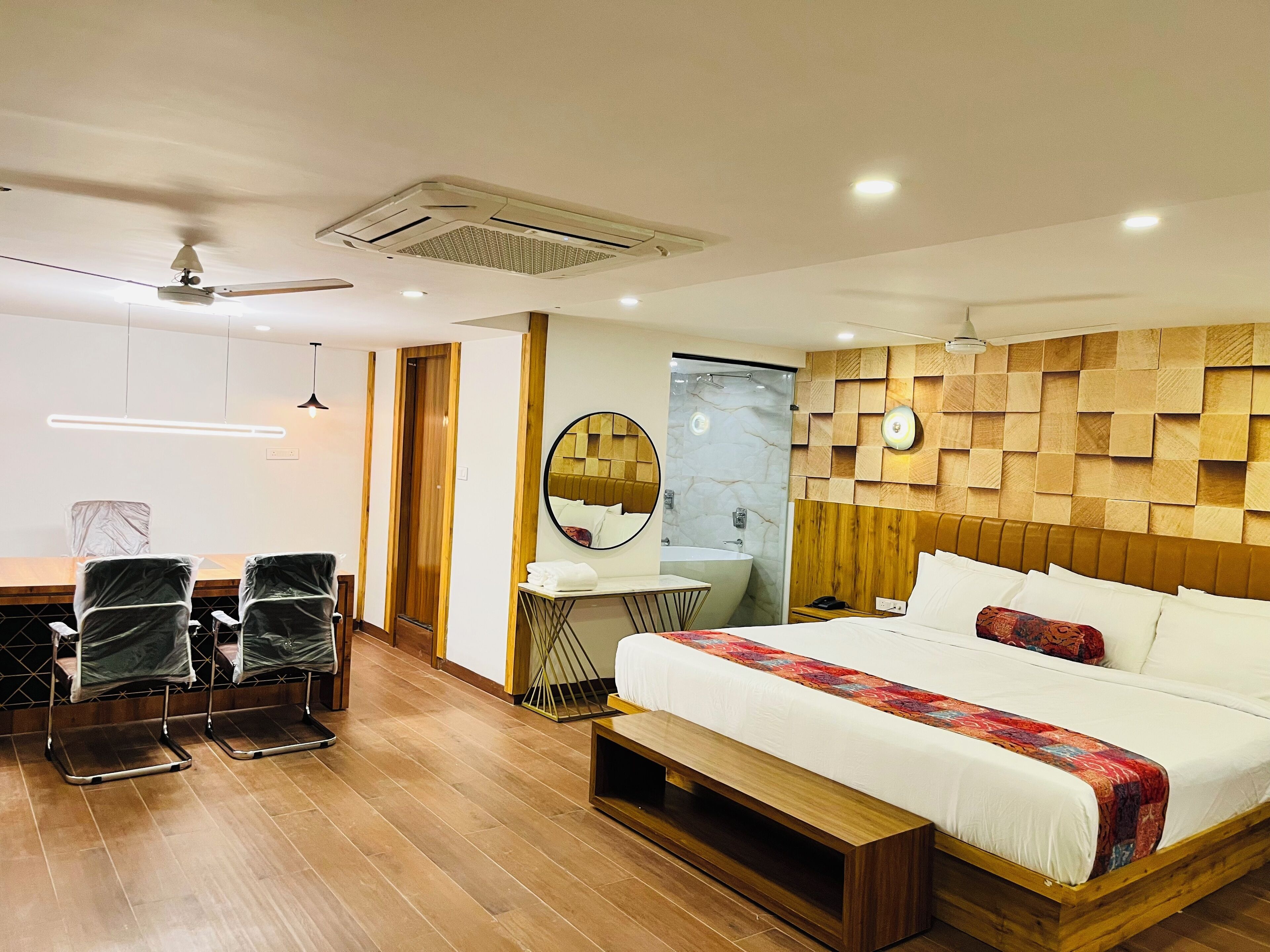 A modern hotel room with a wooden floor, a large bed with a colorful blanket, a wooden headboard, a round mirror, a wooden wall panel, a small table with a mirror, a ceiling fan, and a bathroom with a bathtub and a shower.