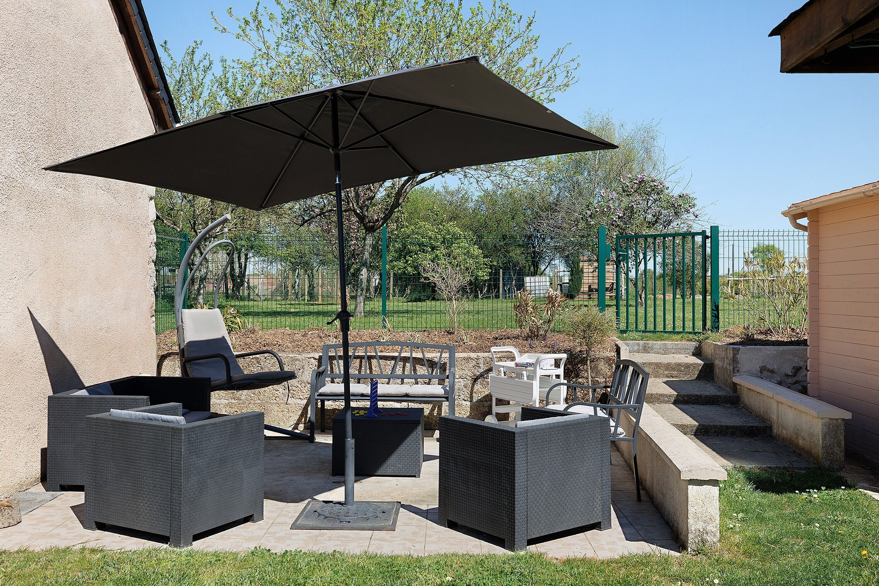 Een terras buiten met wicker meubilair, een grote parasol en een groene hek op de achtergrond.