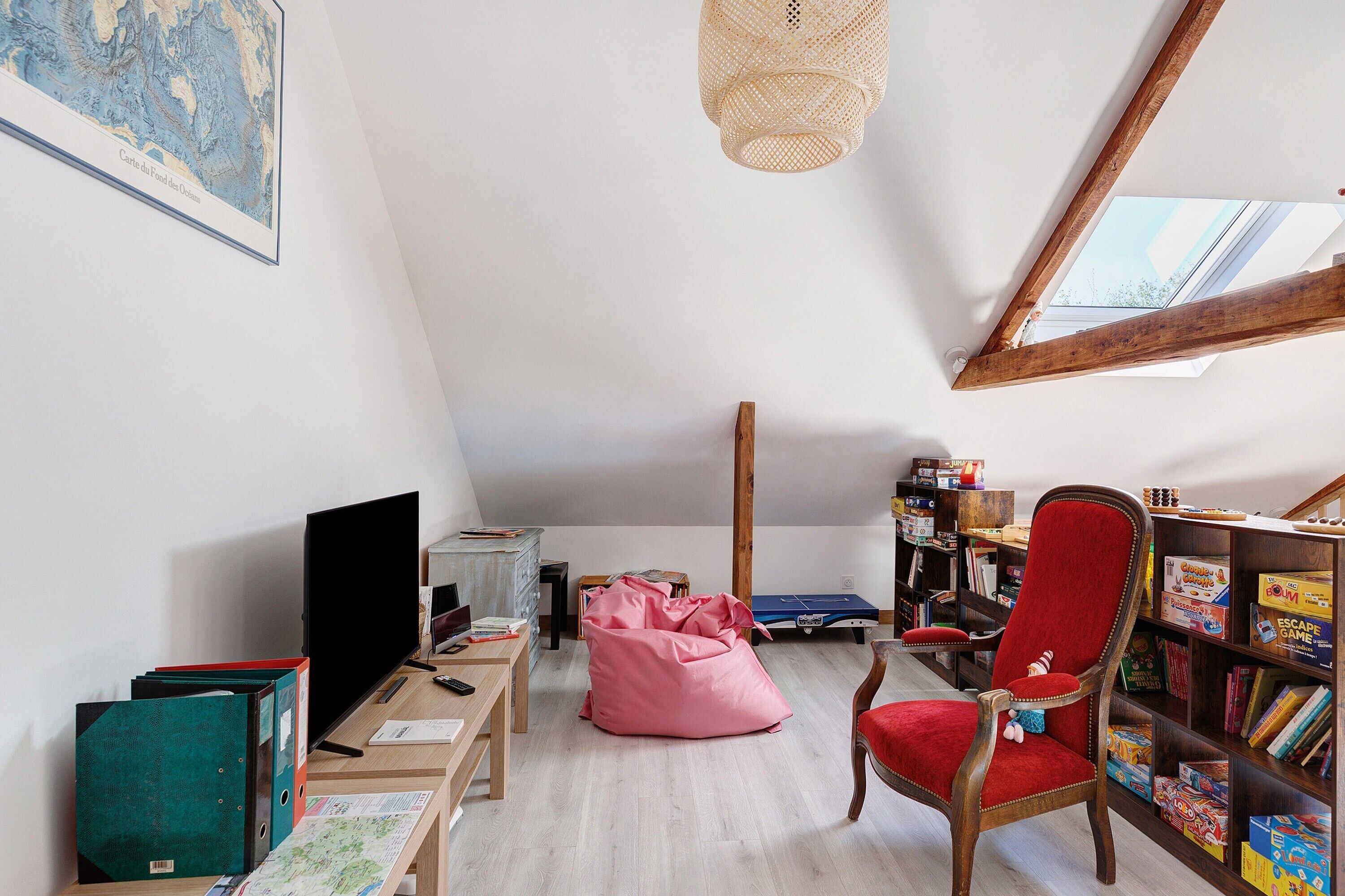 Una habitación con techo inclinado, una estantería, una silla roja, un sillón de saco y un escritorio con computadora.