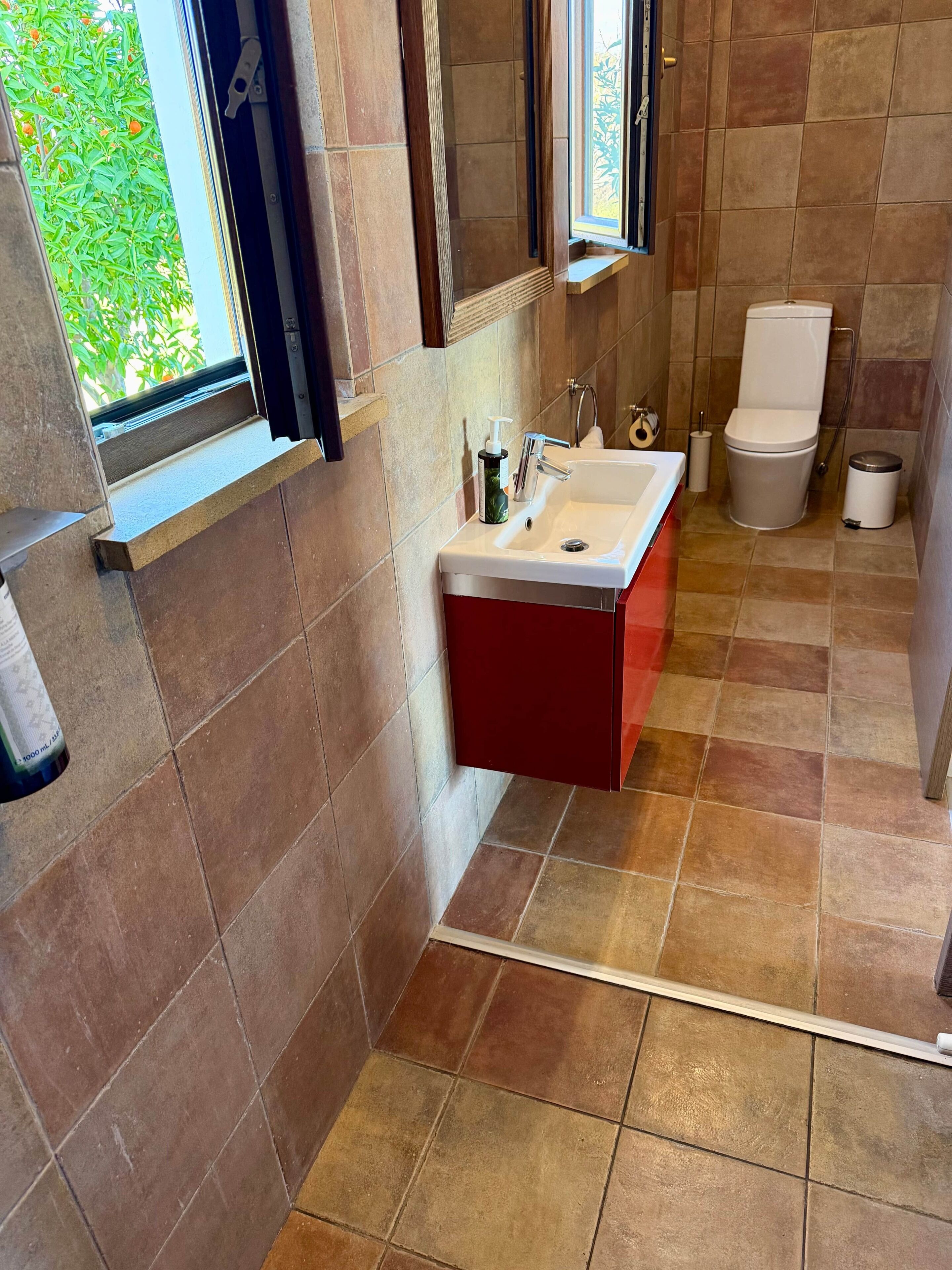 A bathroom with a window, a sink, a toilet, and a trash bin.