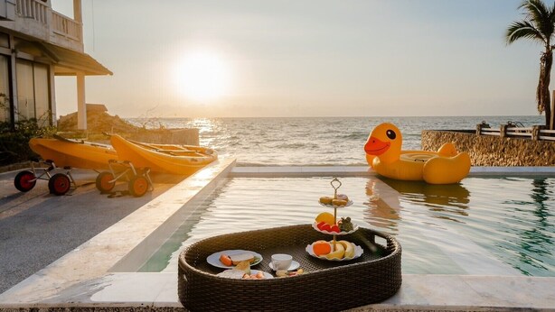 Entorn de piscina amb vista a l'oceà banyat per la llum del sol, una safata de vimet amb menjar i ànecs inflables grocs.