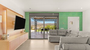 En moderne stue med en grå sofa, et fladskærms-tv på en trækommode og en skydedør, der fører ud til en udendørs terrasse med udsigt over bakkerne.