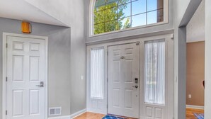 A spacious entryway with a high ceiling, two white doors, a large window with a view of trees, and a chandelier.