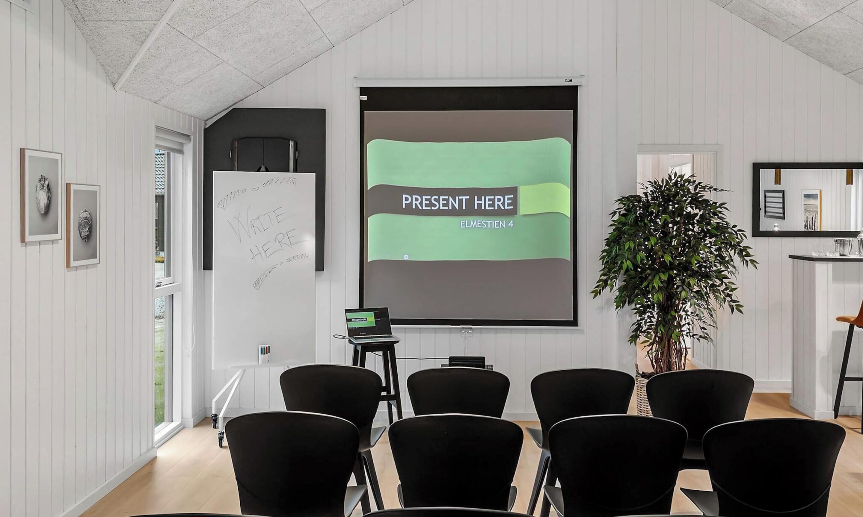 Ein Konferenzraum mit Projektionsschirm, schwarzen Stühlen, einer Whiteboard-Tafel und einem Blumentopf.