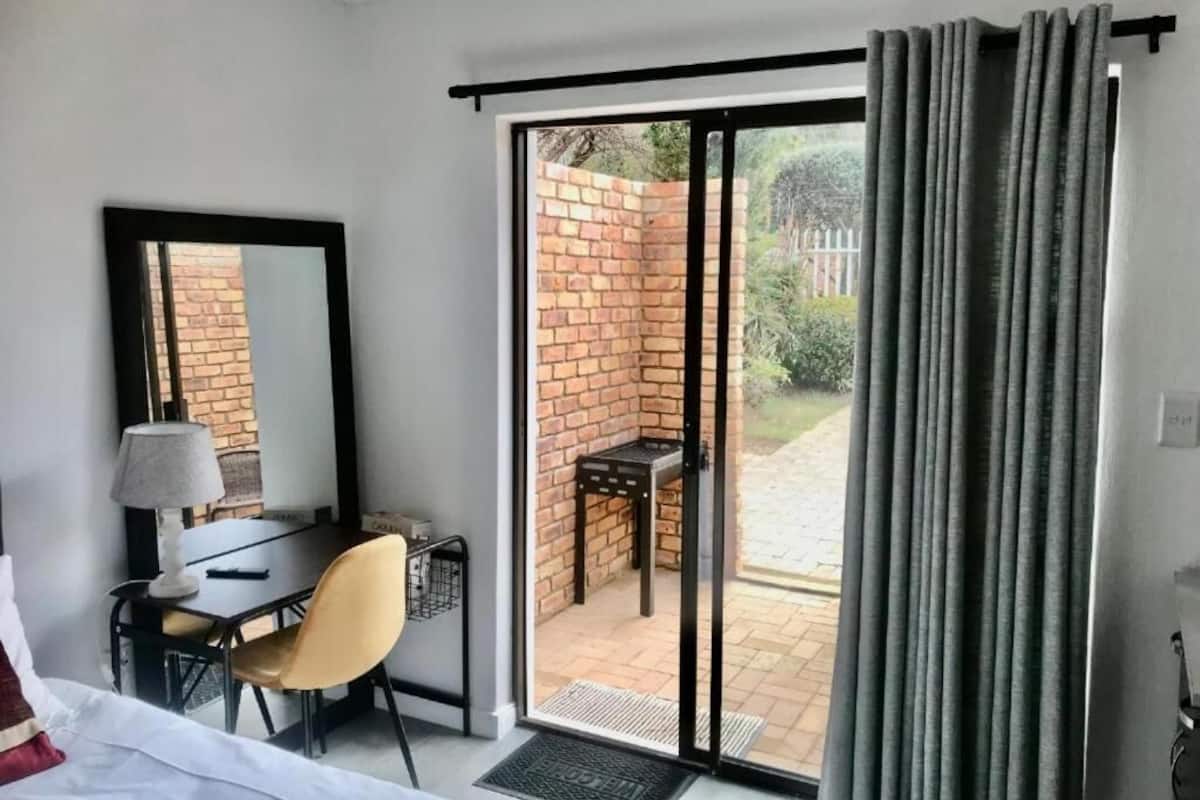 Una habitación de hotel con una cama, un escritorio con una silla, una lámpara y un ventanal corredero que da a un patio exterior con pared de ladrillo y una mesa.