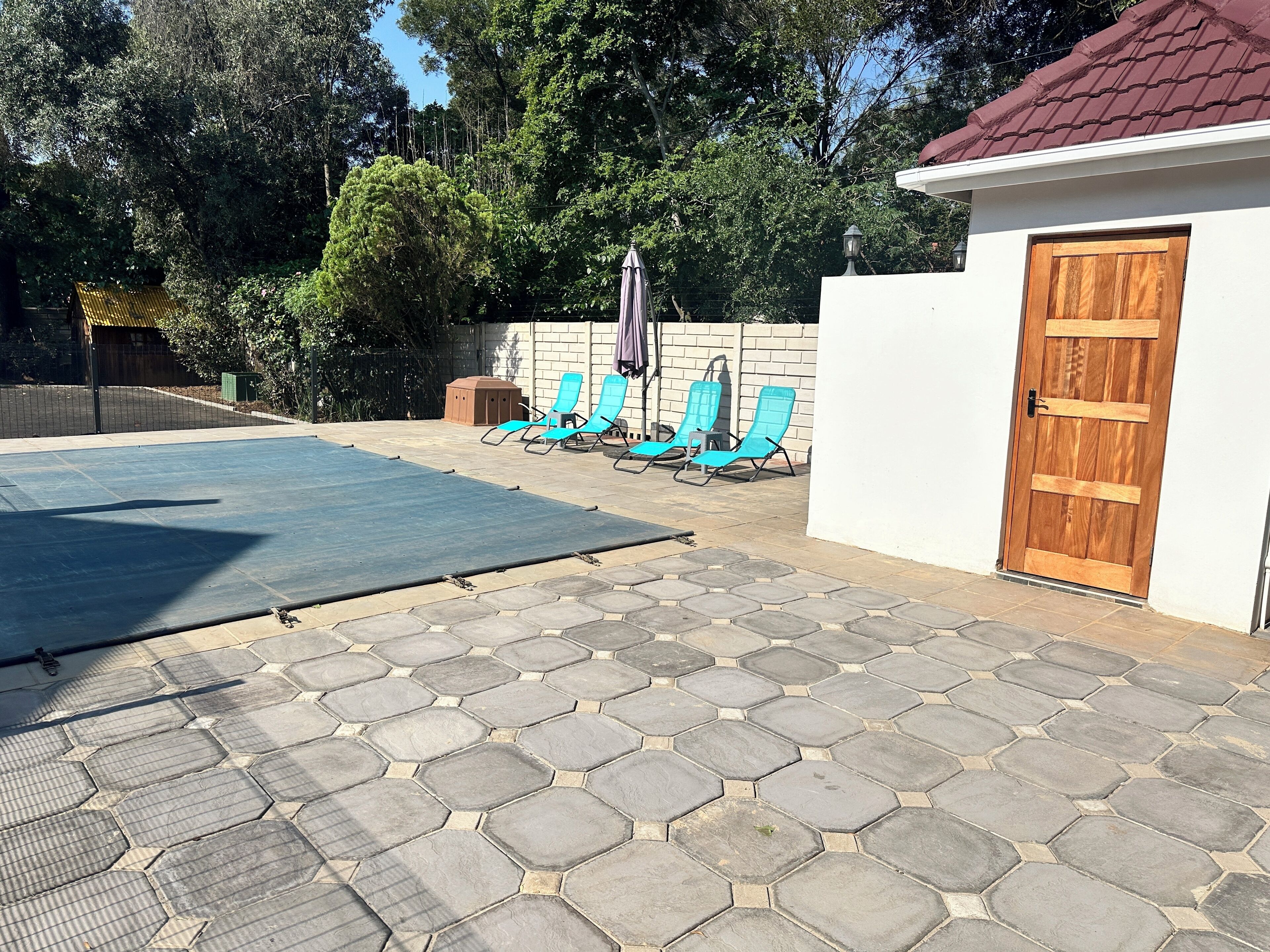 Et overdækket udendørsområde med swimmingpool, sekskantet stenbelægning og blå liggestole.