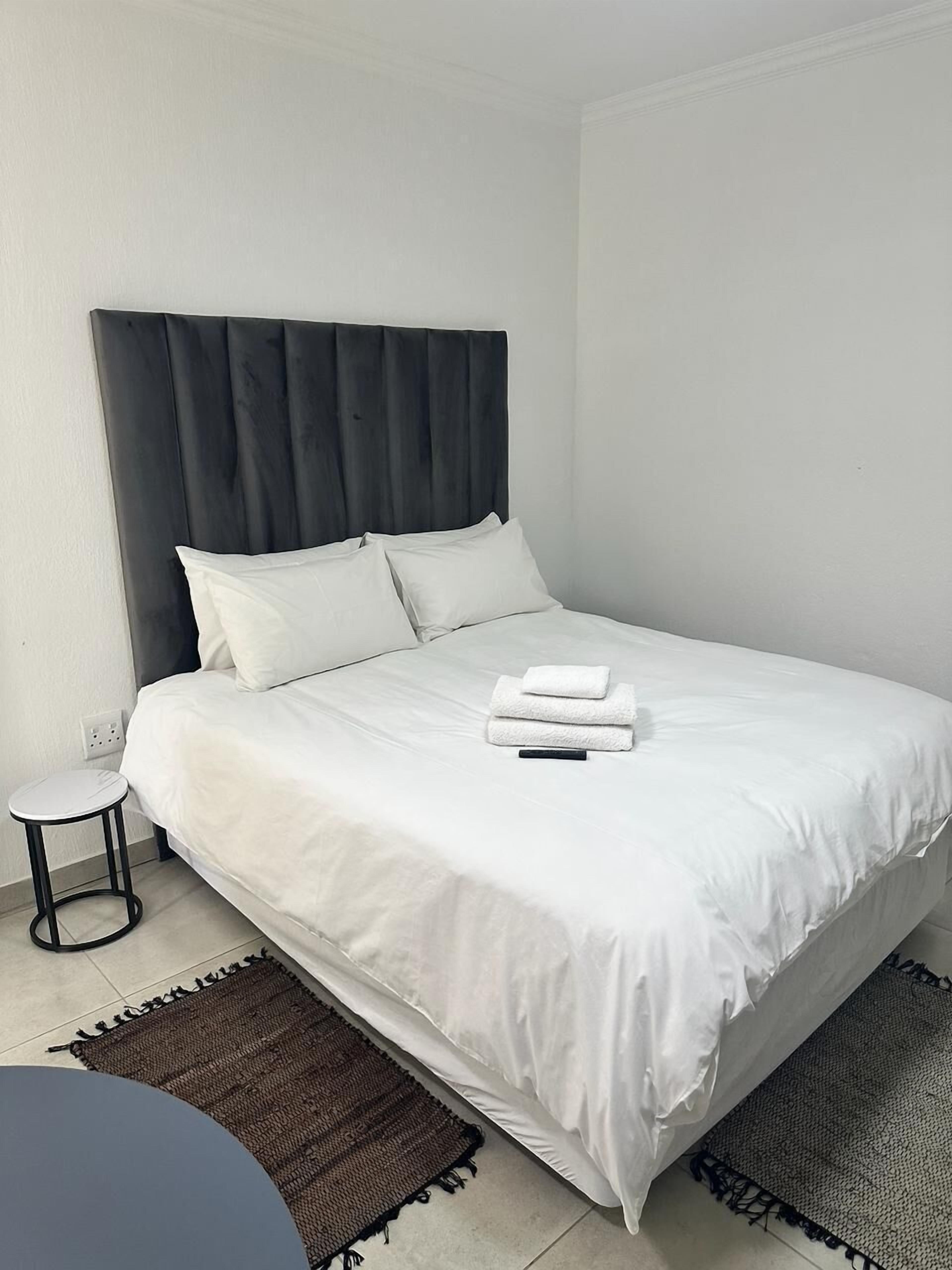 Een netjes opgemaakt bed met witte lakens en kussens, een klein rond tafeltje ernaast, en twee bruine vloerkleden.