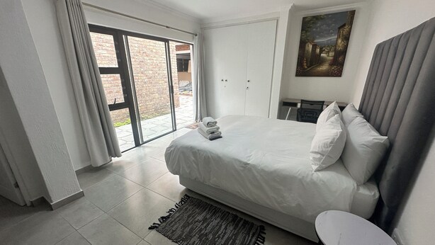 A hotel room with a large bed, a small table, a chair, a painting on the wall, and a sliding glass door leading to a courtyard.