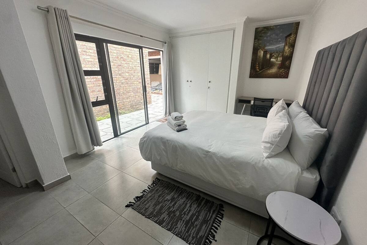 Habitación de hotel con una cama grande, una mesita pequeña, una silla, un cuadro en la pared y un ventanal corredero que da a un espacio exterior.