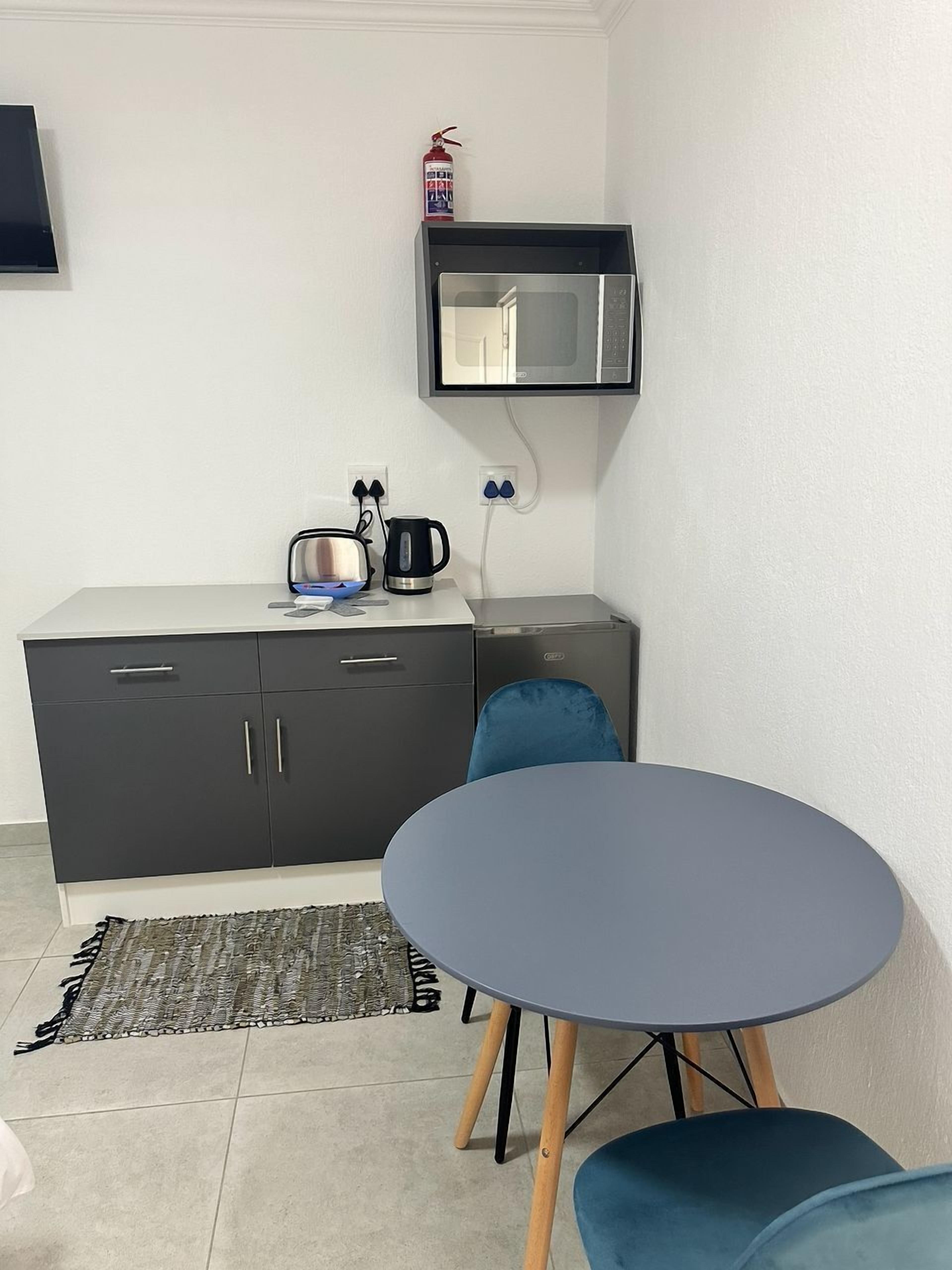 A compact kitchenette with a microwave, kettle, and toaster on a white countertop. There is a blue chair and a small table in the foreground.