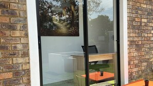 A modern building with a large glass door, brick walls, and a small outdoor seating area with a table and chairs.
