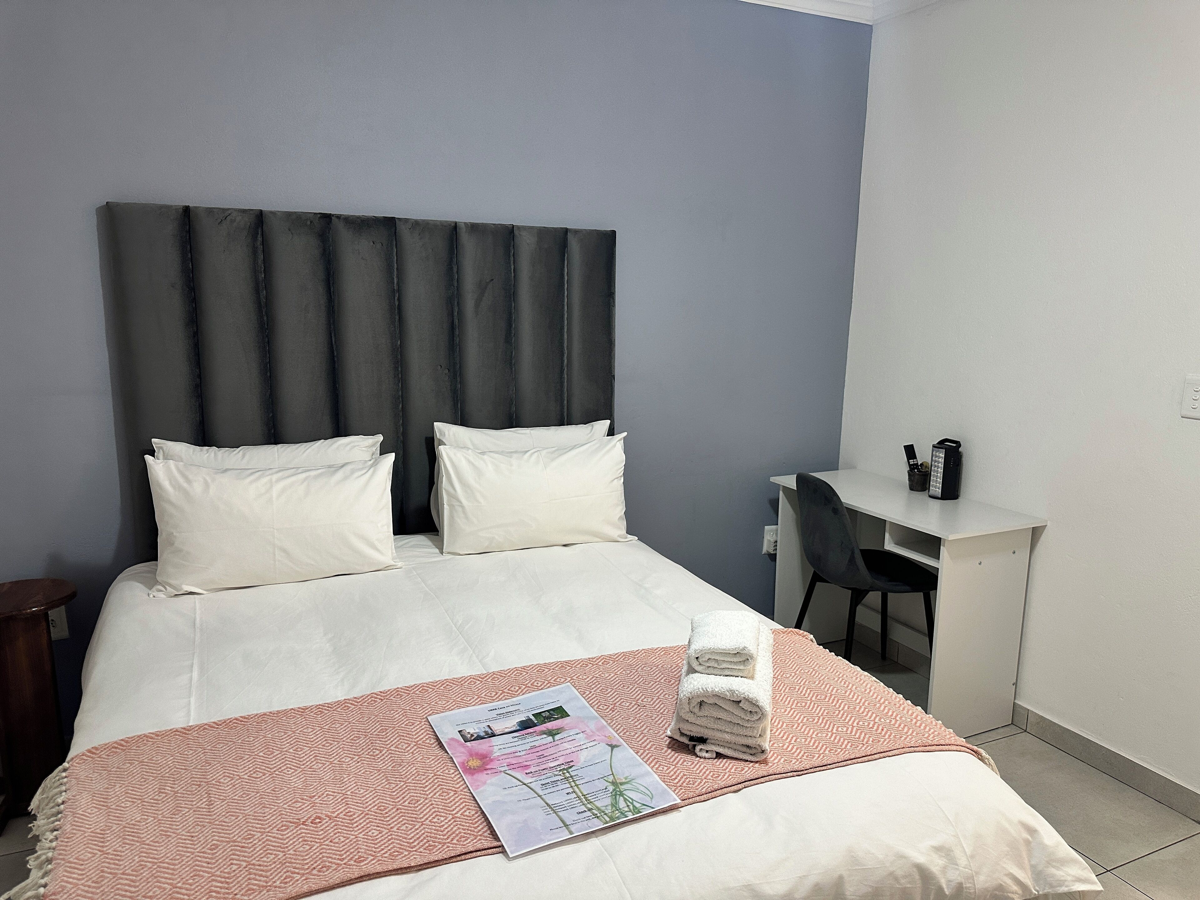 A neatly made bed with white linens and a pink patterned throw, a small desk with a chair, and a wall-mounted phone.