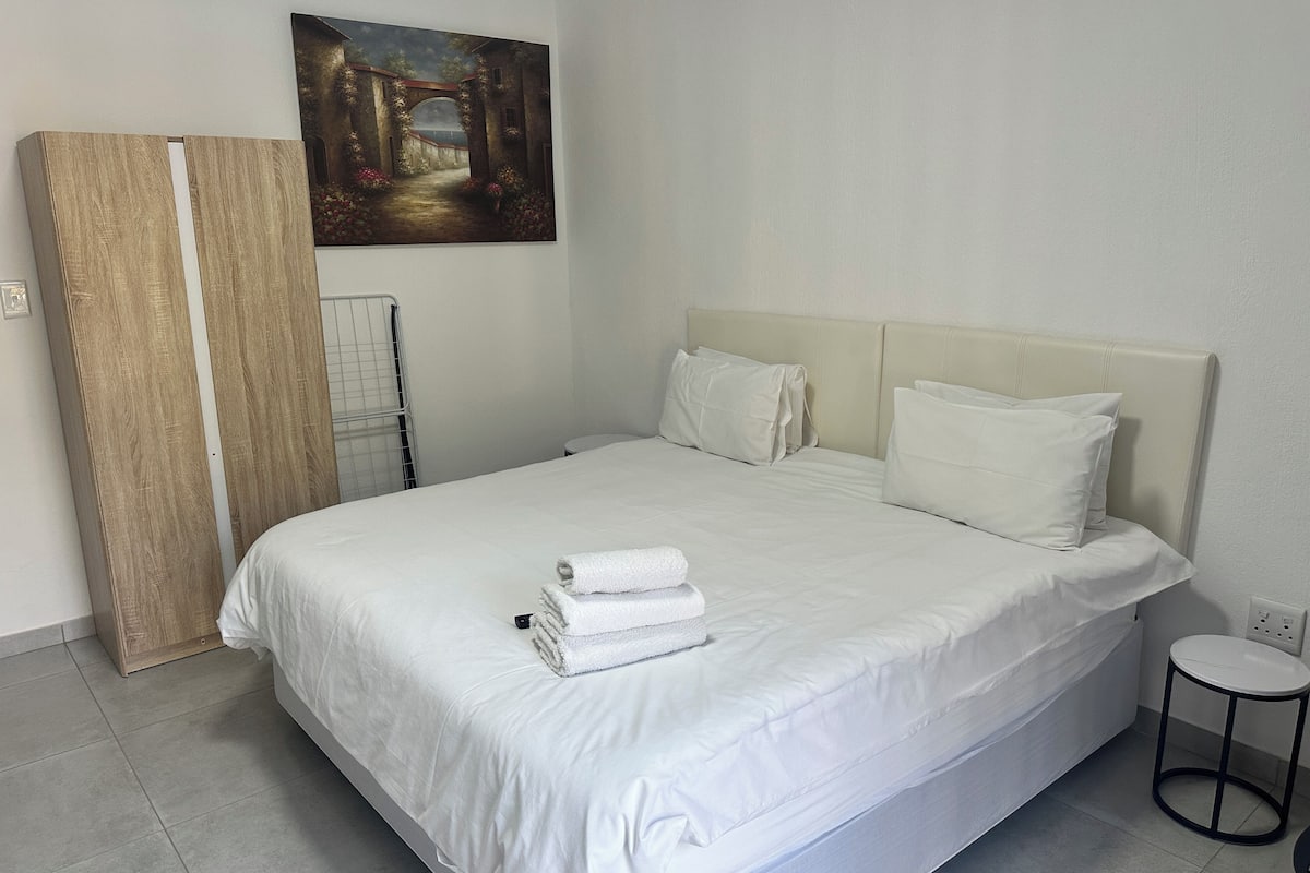 Una cama bien hecha con sábanas y almohadas blancas, un cuadro en la pared y un armario de madera.