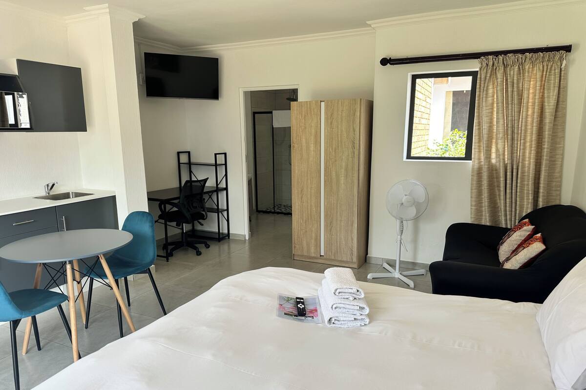 Habitación de hotel con cama, escritorio, silla, televisor, ventilador y ventana con cortinas.