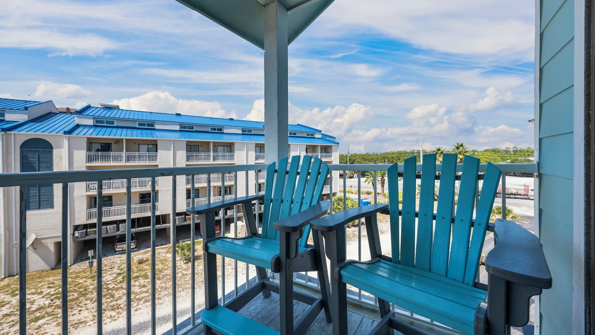 A balcony with two blue Adirondack chairs, a metal railing, and a view of a building with blue roofs and multiple balconies.