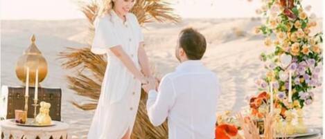 Escena de playa con una pareja tomándose de las manos bajo un arco floral, rodeada de objetos decorativos y un farol.
