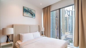 A modern hotel room with a large bed, two bedside lamps, a city view, and a painting on the wall. - Luxury 1BR in Marina Gate with Infinity Pool (Dubai)