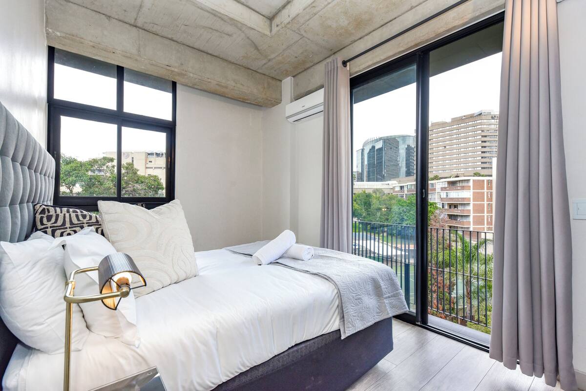 A modern bedroom with a large bed, a bedside lamp, a view of the city through a balcony.