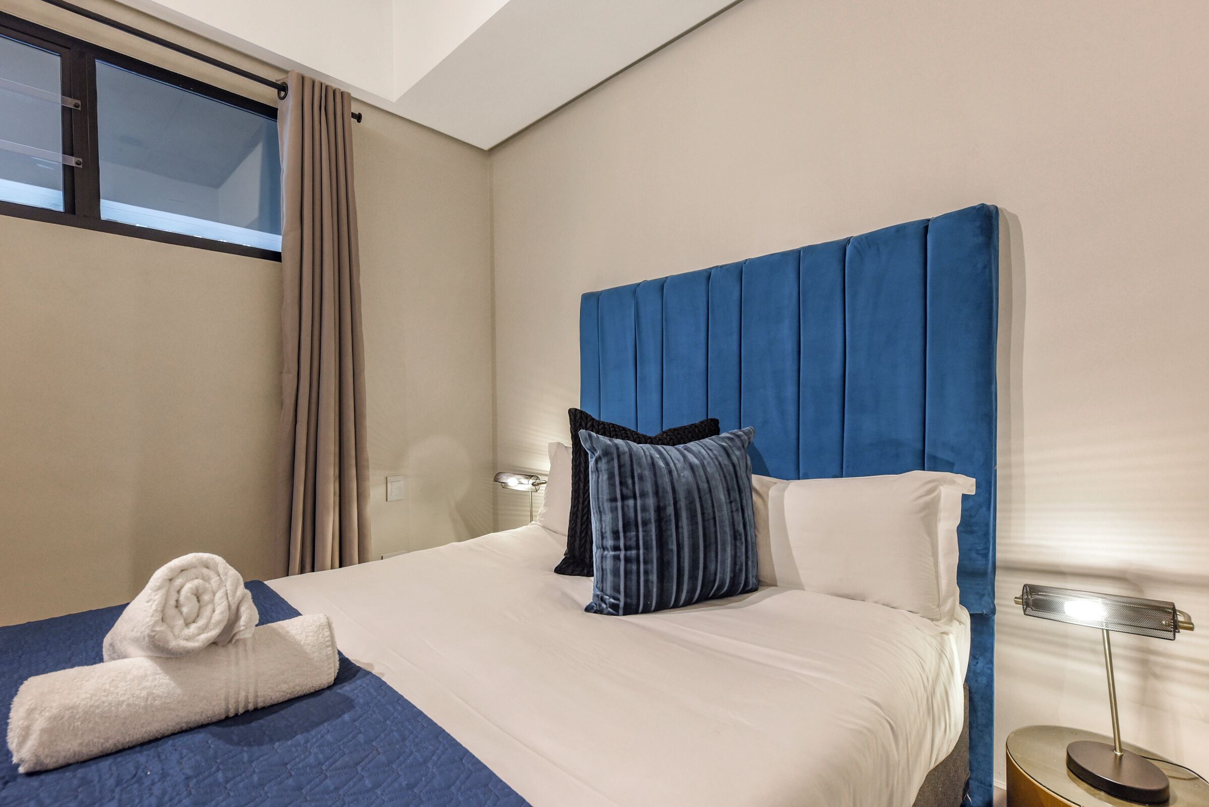 A hotel room with a bed, a nightstand, a lamp, a window with curtains, and a towel on the bed.