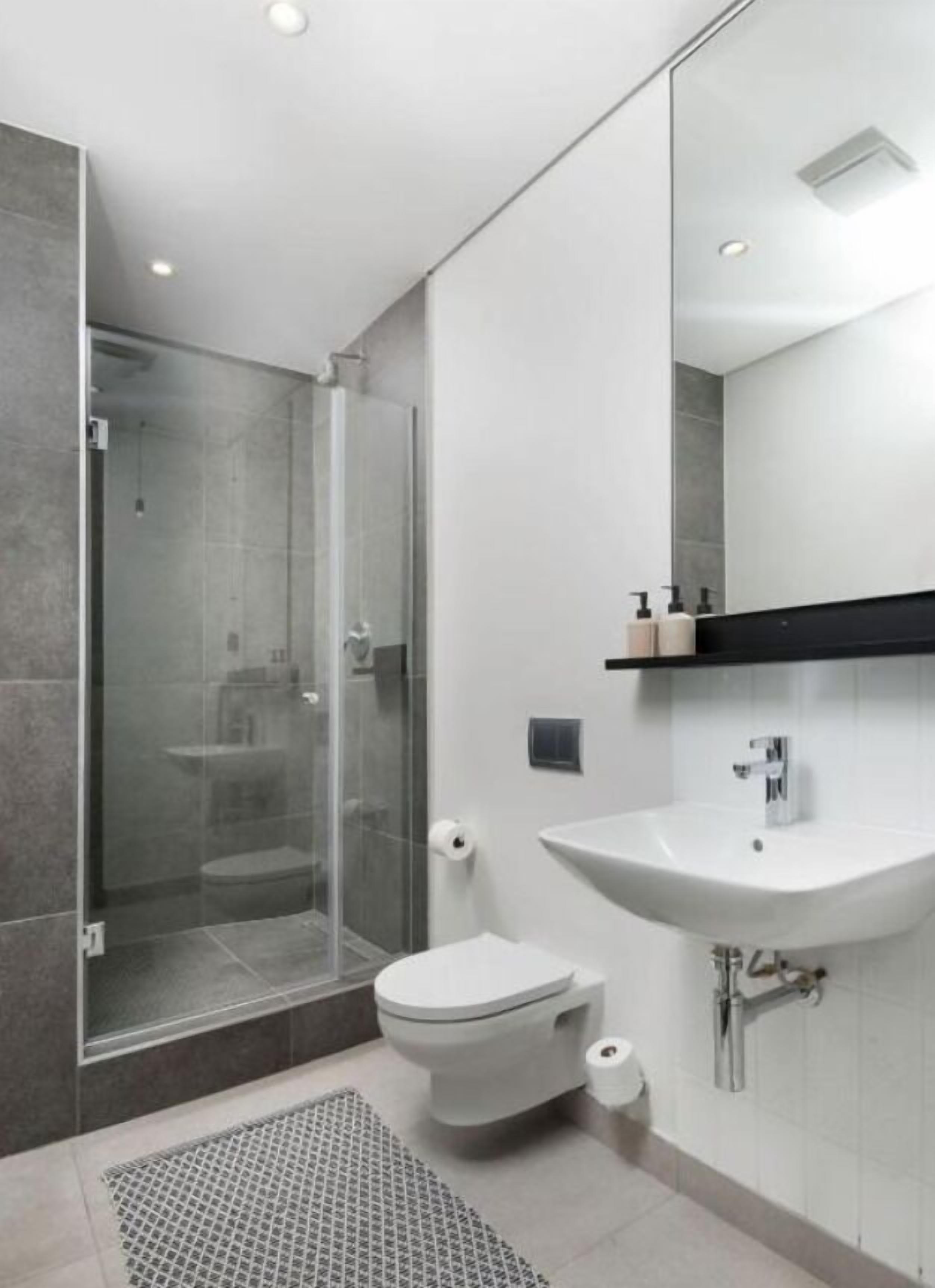 A modern bathroom with a glass shower enclosure, a white toilet, a sink with a mirror above it, and a small shelf with toiletries.