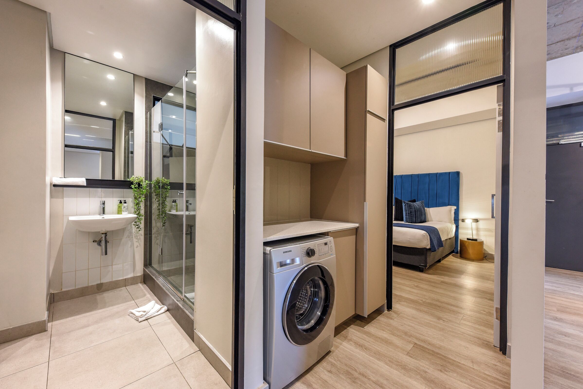 A compact hotel room with a washing machine, a sink, a bed, and a glass shower enclosure.