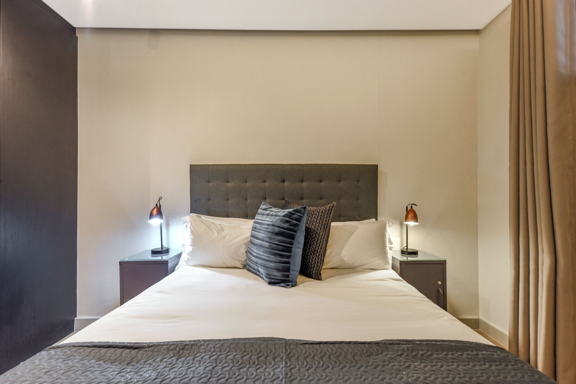 A neatly made bed with a tufted headboard, two bedside lamps, and a dark-colored throw blanket.