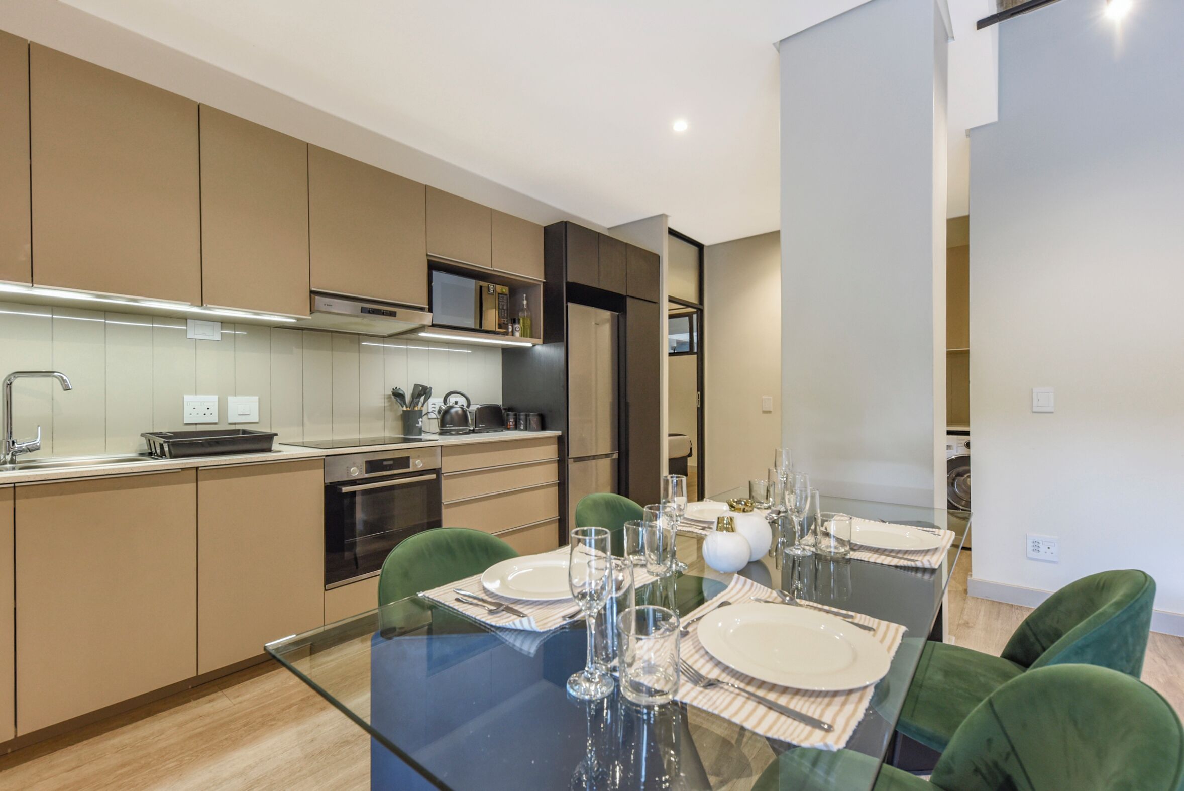 A modern kitchen with a dining area, including a glass table set for four, green chairs, and a built-in oven.