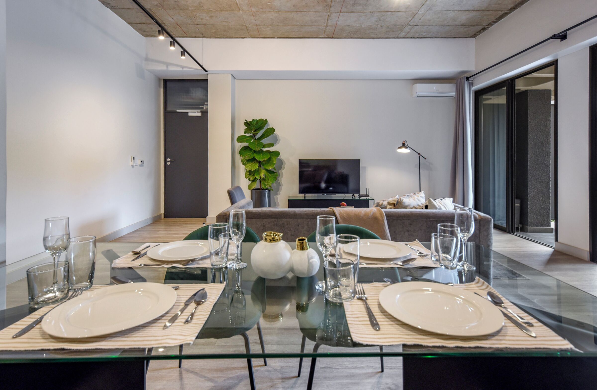 Een moderne eetruimte met een glazen tafel voor vier personen, een bank, een televisie en een plant op de achtergrond.